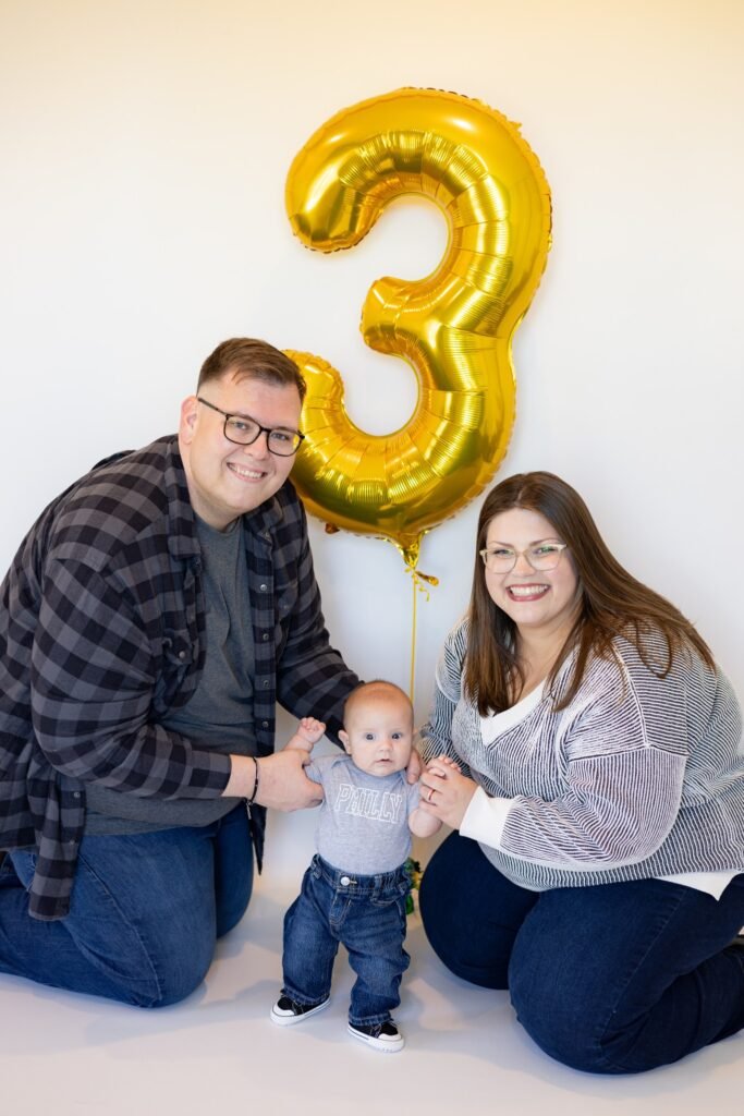 couple posing with their child in front of a balloon shaped like the number 3
