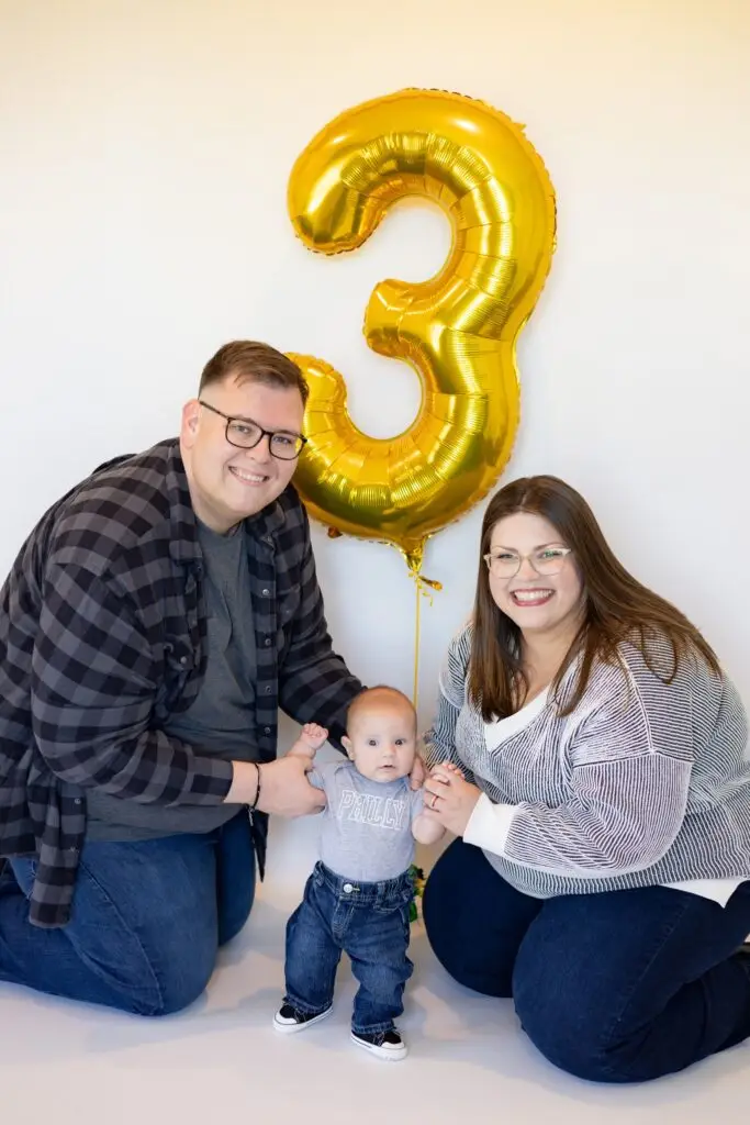couple posing with their child in front of a balloon shaped like the number 3